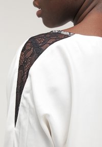 Vit blus med en svarts lace-accent på axeln. Slätt tyg med en avslappnad passform och en subtil glans. Utförlig axeldesign.