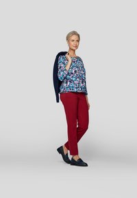 Hemd mit floralem Muster in Blau und dreiviertel Ärmeln, rote Hose und dunkelblaue Slipper, navyfarbene Jacke über die Schulter gehalten.