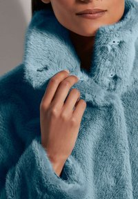 Hellblauer Kunstpelzmantel mit hohem Kragen, ausgestattet mit Knöpfen und weicher Textur, getragen mit einer Hand, die den Kragen berührt, um Details zu zeigen.