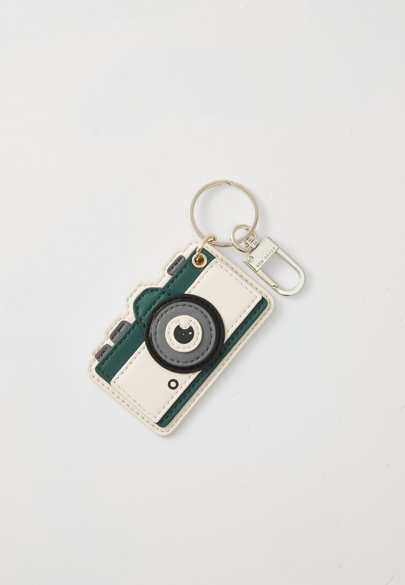 Porte-clés conçu comme un appareil photo, avec un extérieur blanc et vert, un détail de lentille noire ronde, un matériel circulaire et un attachement en métal pour la clé.
