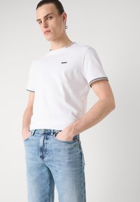 Hvit bomulls t-skjorte med svart logo, stripete mansjetter og rund hals, sammen med lyseblå denimjeans. Modellen har hendene i lommene.