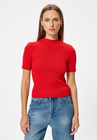 Top rosso a maniche corte con un design a collo alto. Silhouette aderente, texture liscia. Abbinato a jeans blu a vita alta.