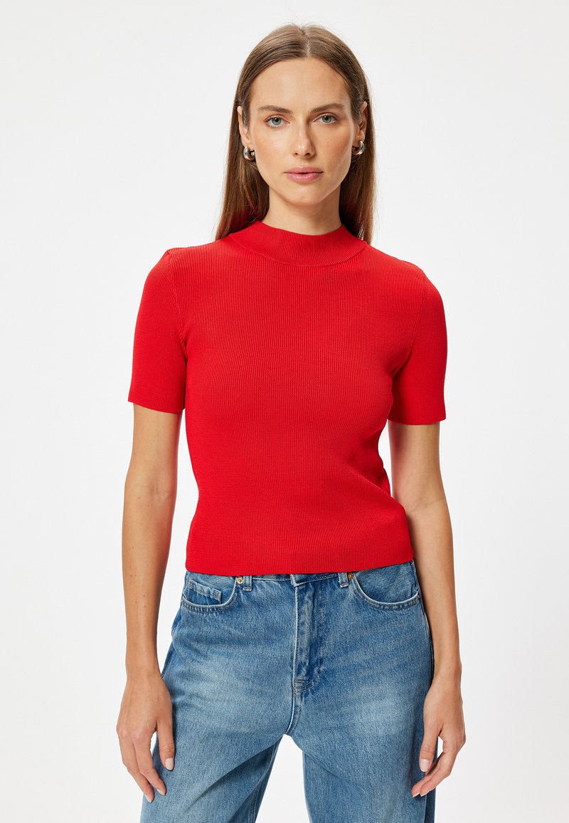 Top rosso a maniche corte con un design a collo alto. Silhouette aderente, texture liscia. Abbinato a jeans blu a vita alta.