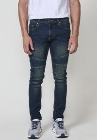 Jeans de mezclilla azul oscuro con un corte ajustado, que presentan detalles acanalados en las rodillas y costuras en contraste. Llevados con zapatillas deportivas grises.