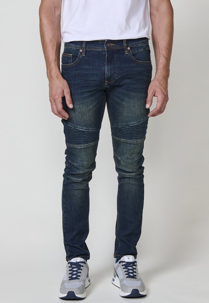 Jeans de mezclilla azul oscuro con un corte ajustado, que presentan detalles acanalados en las rodillas y costuras en contraste. Llevados con zapatillas deportivas grises.