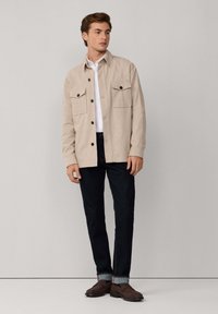 Camisa chaqueta beige de botones con dos bolsillos en el pecho, llevada sobre una camisa blanca. Jeans oscuros y zapatos marrones completan el conjunto.