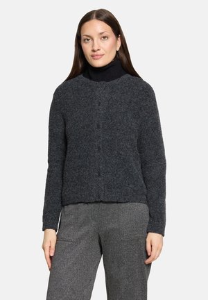 Woman with long dark hair wearing a dark gray buttoned sweater over a black turtleneck and gray pants, standing against a plain background.