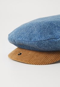 Boné plano com parte superior em denim azul e aba em corduroy bege. A aba apresenta um detalhe de botão de metal preto. O tecido texturizado contrasta entre as seções.