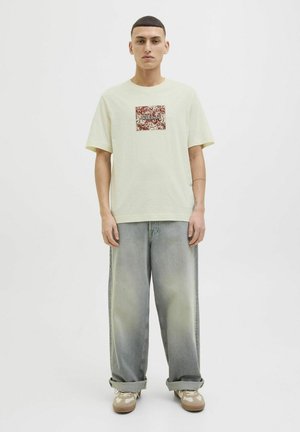 Light yellow short-sleeve shirt features a square maroon floral patch. Worn with faded light blue oversized jeans and beige sneakers.