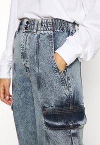 Pantaloni cargo in denim di colore azzurro chiaro. Presentano una vita alta, due grandi tasche laterali e una texture sbiadita con accenti scuri a contrasto.