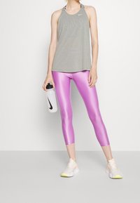 Camiseta deportiva gris de Nike con diseño racerback, combinada con leggings rosas brillantes. La modelo sostiene una botella de agua transparente y lleva zapatillas blancas con detalles amarillos.