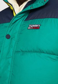 Chaqueta acolchada verde con una sección superior azul marino. Presenta un botón de presión negro y el logo bordado "TOMMY JEANS" en rojo y blanco.