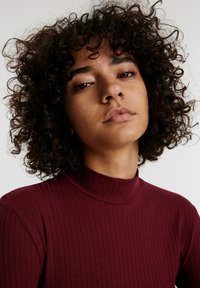 Burgunderfarbener gerippter Rollkragenpullover mit figurbetontem Design. Verfügt über einen hohen Kragen und strukturierten Stoff. Lockiges, dunkles Haar sichtbar.