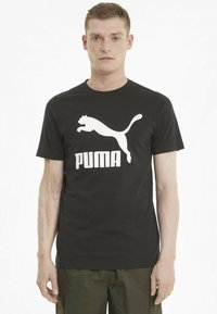 Must must T-särk, millel on ees suur valge hüppav puma logo ja julge "PUMA" tekst. Lihtne ümar kaeluse disain.