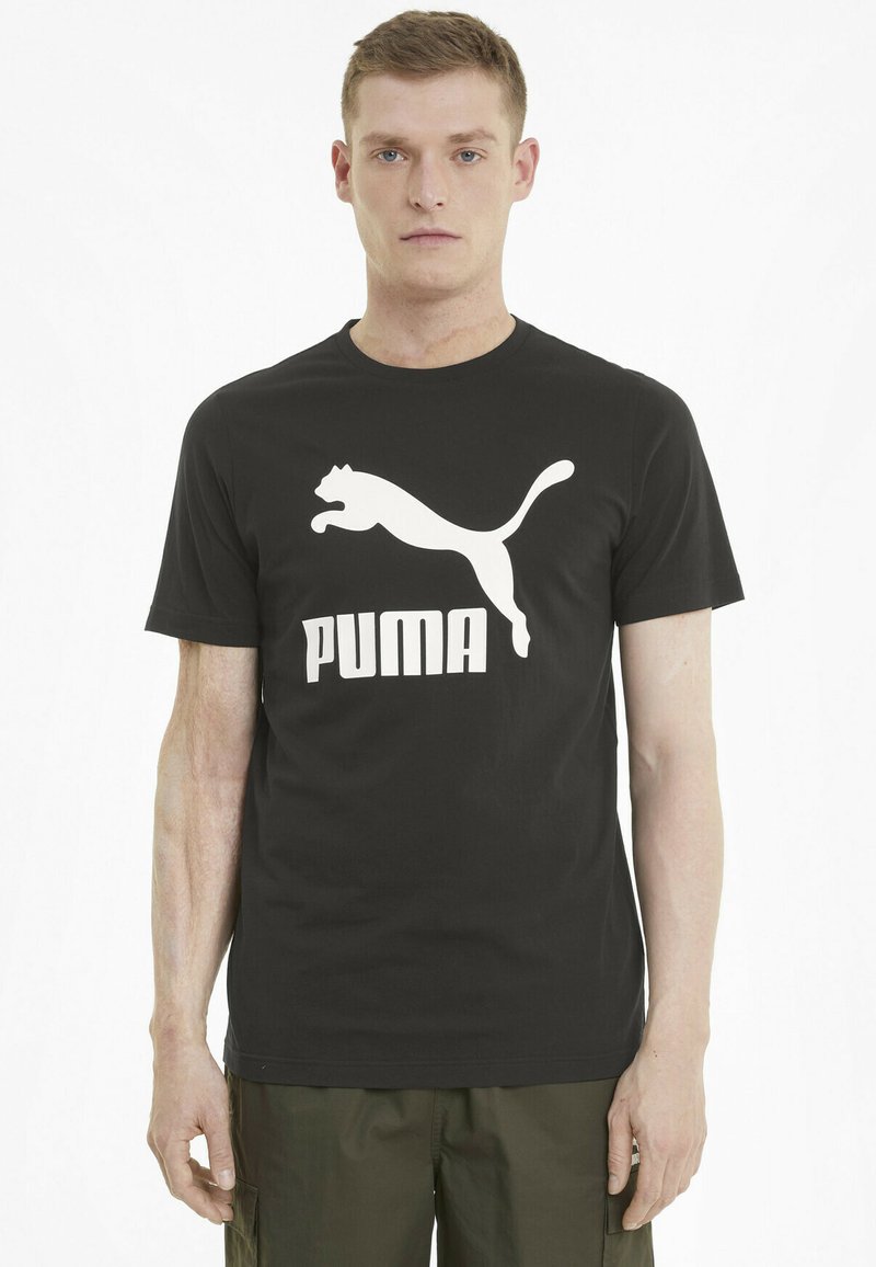 Must must T-särk, millel on ees suur valge hüppav puma logo ja julge "PUMA" tekst. Lihtne ümar kaeluse disain.