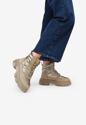 Botas altas beige con acentos metálicos plateados, que presentan un cuello acolchado, una suela gruesa y superficies texturizadas. Llevan cordones con herrajes dorados.