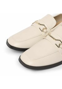 Mocasines de cuero beige con un detalle de cadena dorada. Presentan una textura suave, punta redondeada y una suela de goma negra contrastante con un patrón en zigzag.
