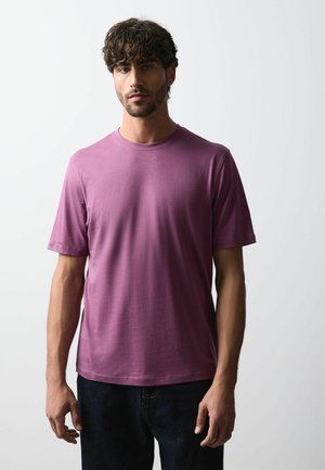 Ung mand med krøllet mørkt hår iført en ensfarvet lilla t-shirt med korte ærmer og mørke bukser mod en ensfarvet lys baggrund.
