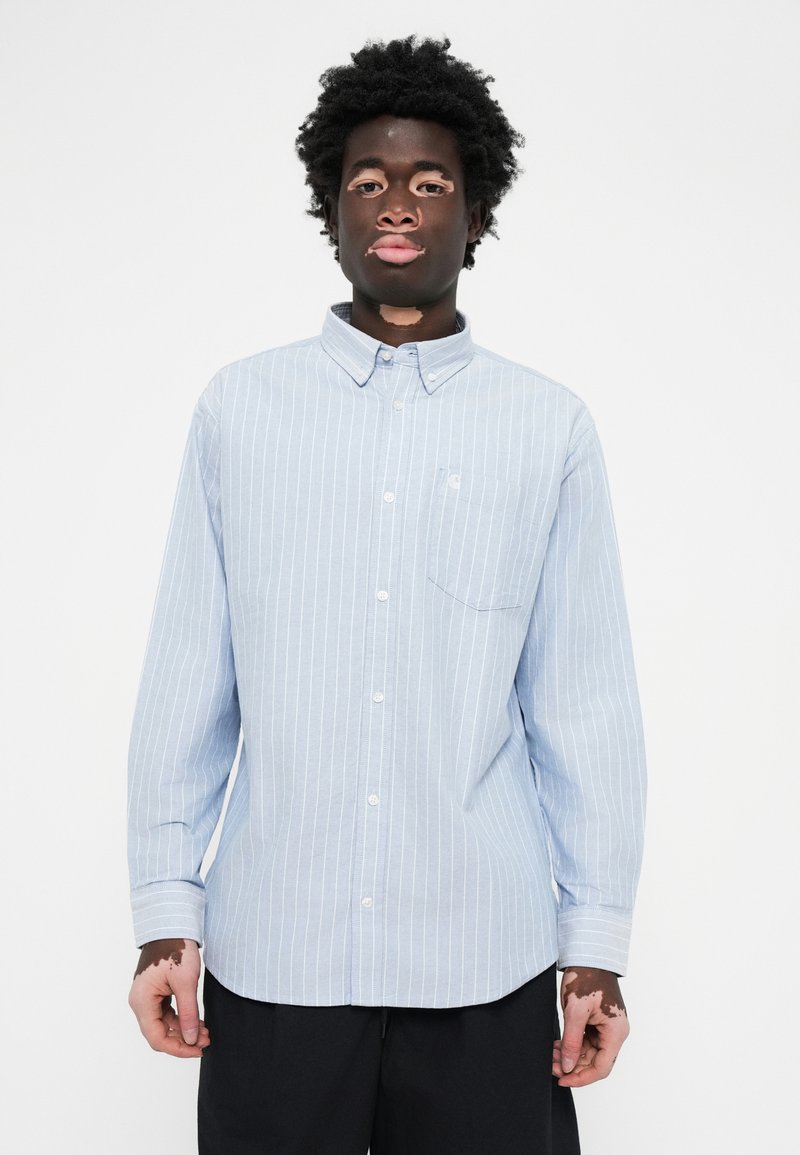 Chemise boutonnée rayée bleu clair avec col, poche poitrine unique et manches longues. Fabriquée en tissu doux avec une texture lisse.
