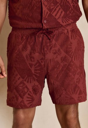 Shorts texturés couleur marron avec un motif de soleil et de main, taille élastique et cordon, portés avec une chemise boutonnée assortie.