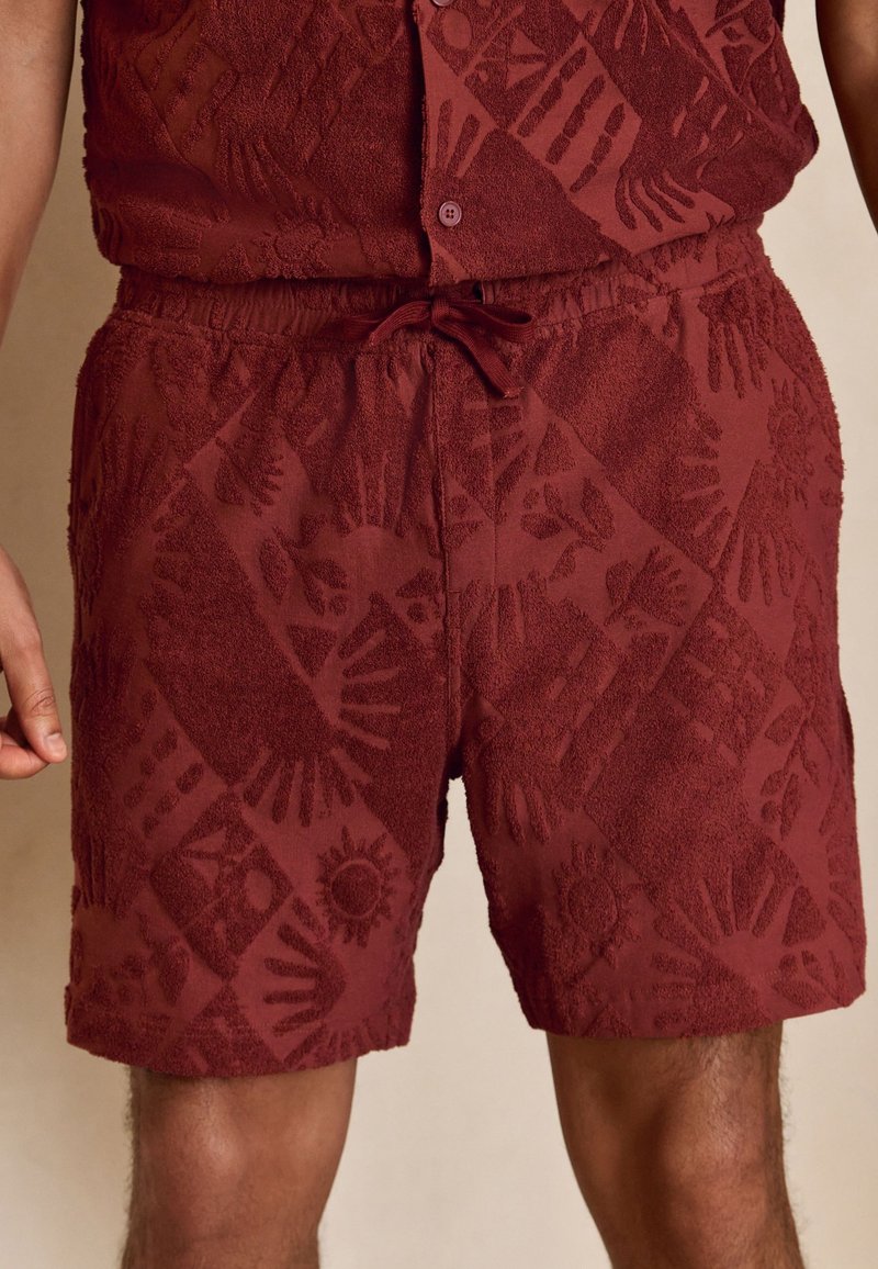 Shorts texturés couleur marron avec un motif de soleil et de main, taille élastique et cordon, portés avec une chemise boutonnée assortie.