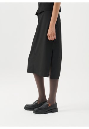 Kvinde iført sort midi-nederdel med sidespalte, mørke gennemsigtige strømpebukser og sorte chunky loafers, stående mod en ensfarvet lys baggrund.