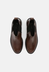 Un par de zapatos marrones de cuero sin cordones con paneles elásticos laterales negros y tiradores, vistos desde arriba sobre un fondo blanco.