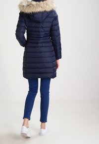 Marineblauwe pufferjas met een bontgevoerde capuchon, gewatteerde structuur, lange mouwen en een aansluitende pasvorm. Gemaakt in combinatie met blauwe spijkerbroeken en witte sneakers.