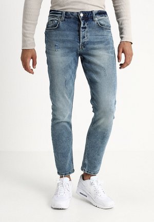 Mand iført lyseblå falmede jeans med let slid og hvide sneakers står foran en ensfarvet hvid baggrund.