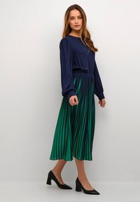 Top navy cu mânecă lungă, cu fustă pliată verde și albastră, având un talie lată. Modelul poartă pantofi cu toc negri.