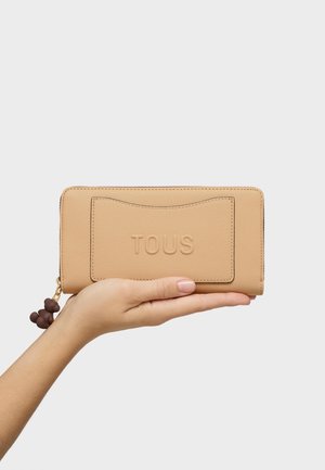 Mano sosteniendo una cartera beige de TOUS con el logo en relieve y un tirador de cremallera en forma de oso marrón sobre un fondo gris claro.