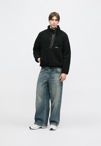 Juodas vilnonis megztinis su ketvirčio užtrauktuku ir krūtinės kišene, derinamas su laisvo kirpimo šviesiai mėlynomis džinsomis ir baltais sportbačiais. Minimalistinio dizaino.