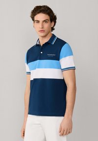 Joven con una camisa polo a rayas en azul, celeste y blanco con el texto "FACONNABLE", combinada con pantalones blancos, sobre un fondo liso.