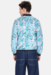 Giacca bomber floreale con una base rosa, caratterizzata da motivi di foglie tropicali blu, colletto e polsini neri. Indossata con pantaloni in denim blu.