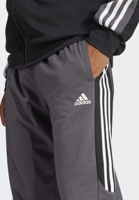 Pantaloni sportivi grigi con logo Adidas bianco, tasche laterali e strisce laterali nere con tre linee verticali bianche. Materiale morbido.