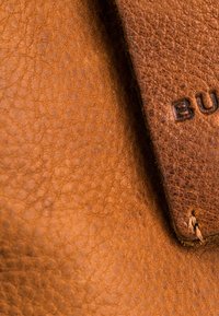 Texture en cuir brun avec une surface finement grainée. Une section étiquetée est visible, avec des lettres dorées en relief et un détail de couture.