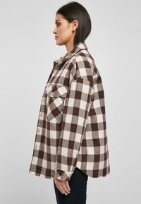 Camisa a cuadros marrón y blanca, estilo abotonado, con cuello y bolsillo en el pecho, que presenta un corte relajado y mangas largas.