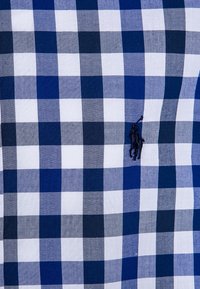 Vzor modré, námořnické a bílé kostkované tkaniny s malým vyšitým tmavě modrým hráčem polo na koni, který drží zvednutou hůl.