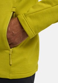 Groene fleece jas met een ritszak en elastische manchetten. Close-up die de gestructureerde stof en het detail van de ritsenpull toont.