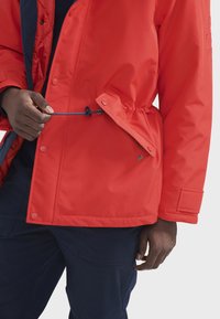 Rote isolierte Jacke mit einer Reißverschlusstasche und verstellbarem Kordelzug in der Taille. Strukturiertes Material und Druckknopfverschluss. Marinefarbene Akzente.