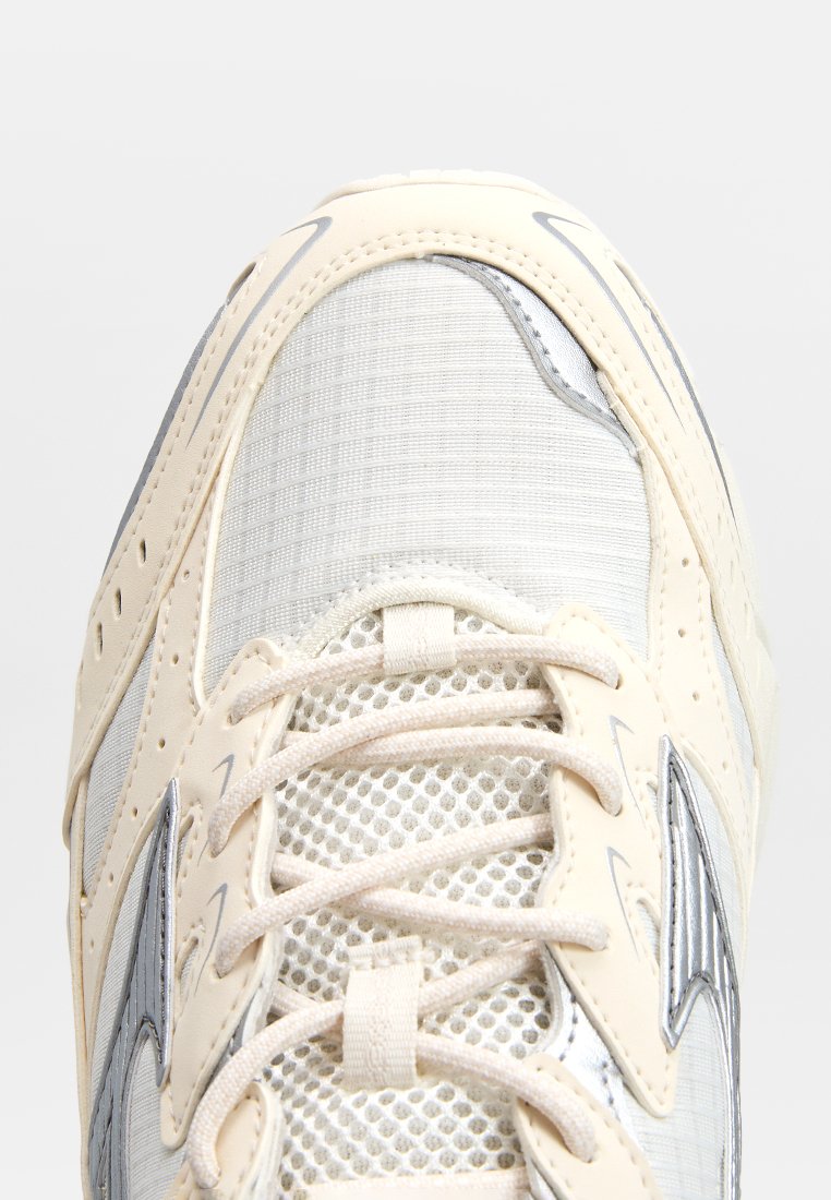 Zapatilla deportiva en color beige claro con parte superior de malla texturizada, detalles en plata y cordones en contraste. Diseño de puntera redondeada y cuello acolchado.