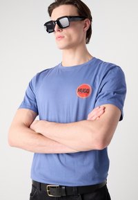 Blue cotton t-shirt with short sleeves, featuring a red circular logo with black text. The model wears black rectangular sunglasses and a black belt.