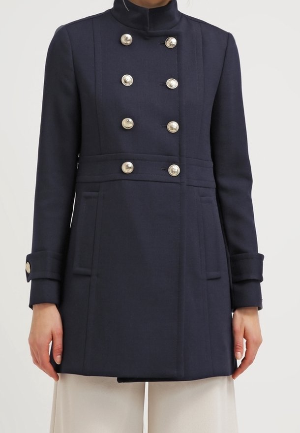 Cappotto navy doppiopetto con colletto alto, bottoni argentati e polsini a campana; presenta due tasche laterali e una vestibilità sartoriale.