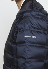 Gros plan d'une personne portant une doudoune bleu marine foncé avec le nom de la marque Michael Kors sur l'épaule arrière.