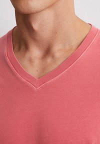 T-shirt à col en V en tissu corail doux. Présente une texture lisse et des coutures délicates autour du col, avec une coupe ajustée.