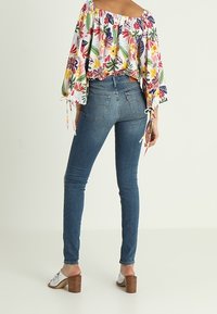 Woman wearing blue skinny jeans, white heeled sandals, and a floral off-shoulder blouse with loose sleeves tied at the wrists.