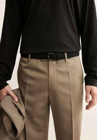 Pantaloni beige su misura con pieghe, abbinati a una camicia nera a maniche lunghe. Cintura nera con fibbia d'argento e giacca in mano.