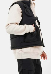 Schwarze gesteppte Weste mit Frontreißverschluss, Seitentaschen und einer Tasche auf der Brust. Getragen über einem hellcremefarbenen Sweatshirt mit Bündchen.