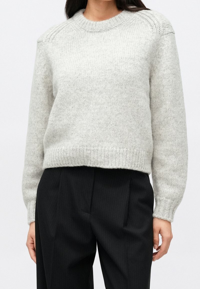 Femme portant un pull en maille gris clair avec des poignets côtelés et un pantalon plissé noir taille haute sur un fond uni.