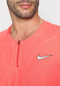 Camisa deportiva con cremallera de Nike en un brillante color coral, que cuenta con una tela texturizada transpirable, mangas cortas y un pequeño logotipo negro en la parte frontal.